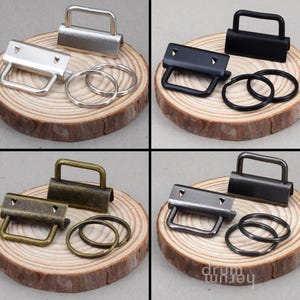 Könnte beinhalten: Vier Sätze Metallbeschläge zum Basteln, jeweils auf einer Holzscheibe. Die Sets umfassen einen rechteckigen Griff, eine rechteckige Basis mit zwei Löchern und zwei Schlüsselringe. Die Beschläge sind in Silber, Schwarz, Bronze und Gunmetal erhältlich.