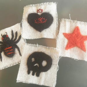 Pode incluir: Quatro retalhos quadrados de tecido bege claro com bordas desfiadas, cada um com um design de feltro de agulha: uma aranha preta com olhos e listras vermelhas, um coração preto com um olho e coroa vermelhos, uma caveira preta e uma estrela laranja.