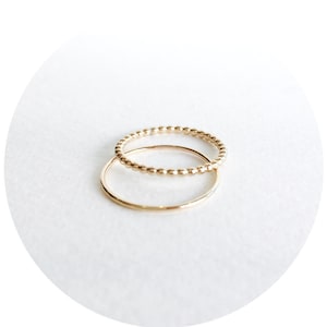 May include: Two gold rings, one with a beaded design and the other a simple band, are stacked on top of each other.
