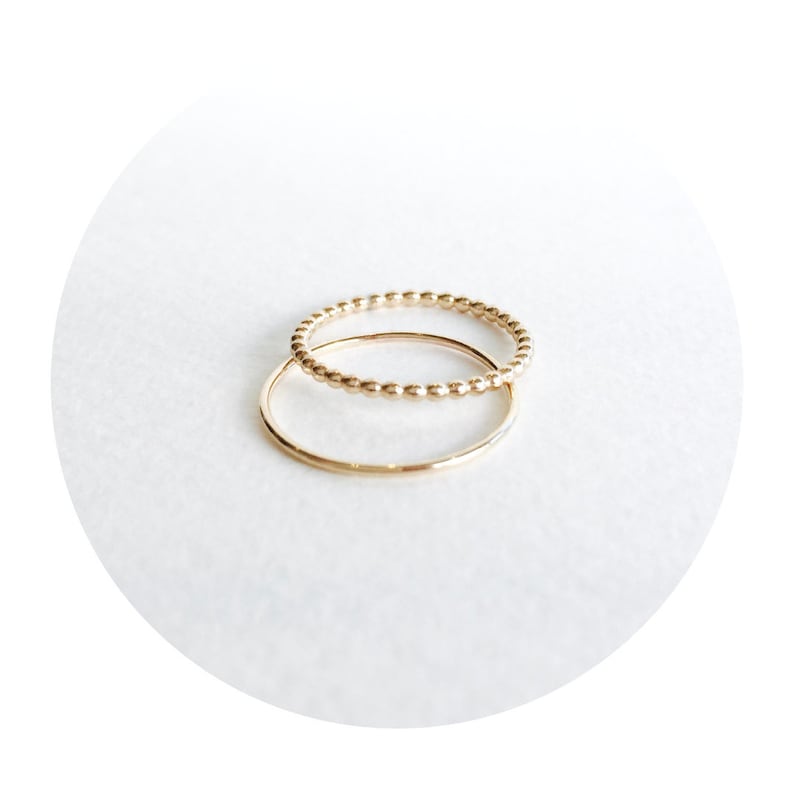May include: Two gold rings, one with a beaded design and the other a simple band, are stacked on top of each other.