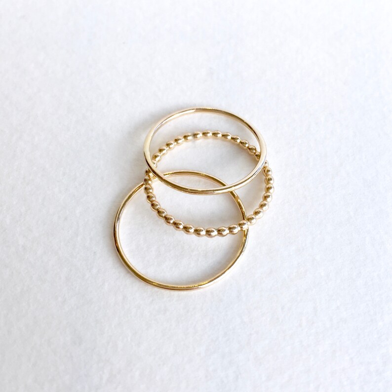 May include: Three gold rings stacked on top of each other. The top ring is a simple band, the middle ring is a beaded band, and the bottom ring is a simple band.