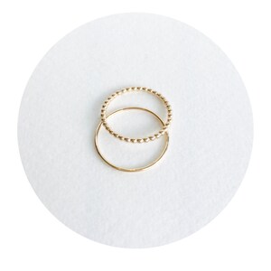 May include: Two gold rings, one with a beaded design and the other a simple band, are stacked on top of each other.