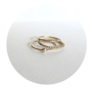 May include: Three gold rings stacked on top of each other. The top ring is a simple band, the middle ring is a beaded band, and the bottom ring is a simple band with a small ball at the end.