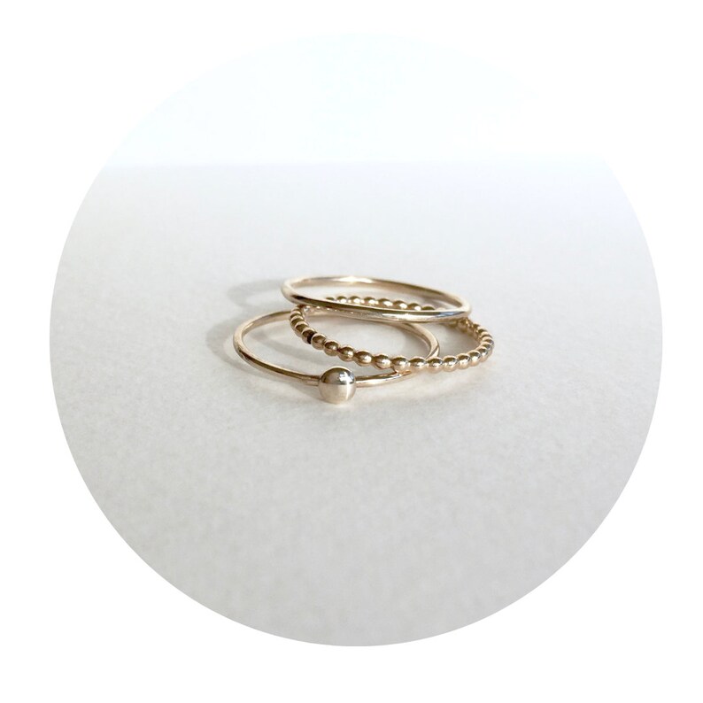 May include: Three gold rings stacked on top of each other. The top ring is a simple band, the middle ring is a beaded band, and the bottom ring is a simple band with a small ball at the end.