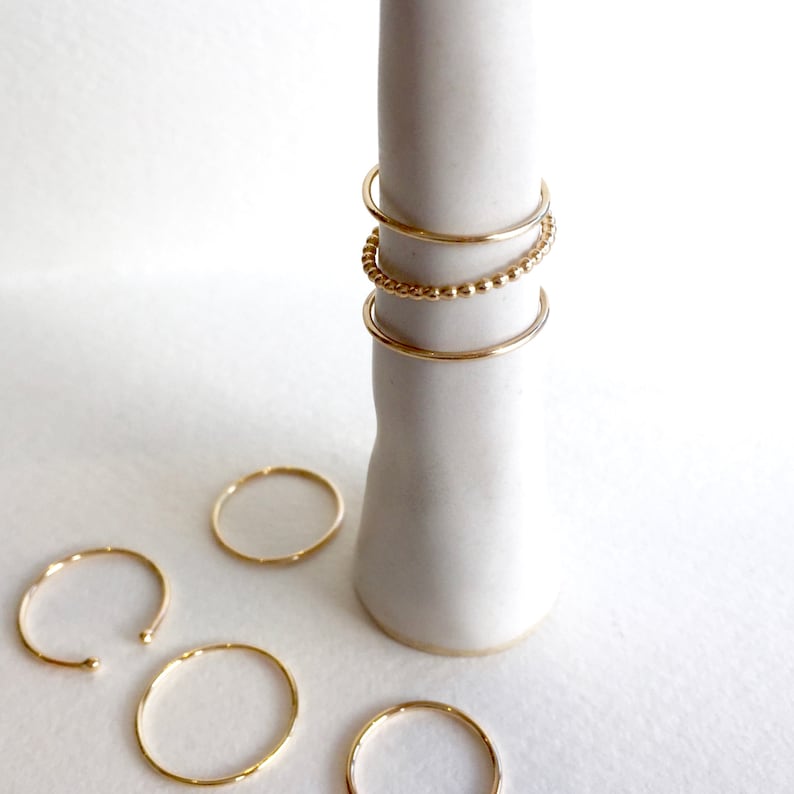 May include: A set of five gold rings, including a beaded ring, on a white ceramic ring holder.
