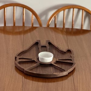 May include: A wooden serving tray shaped like the Millennium Falcon from Star Wars. The tray has a small white ceramic bowl in the center.