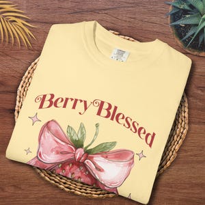 Op de afbeelding: Lichtgeel T-shirt met de tekst "Berry Blessed" in bordeauxrood schrift, vergezeld van een aquarel aardbei illustratie. De aardbei heeft een roze strik en groene bladeren. Het shirt is opgevouwen op een gevlochten mand.