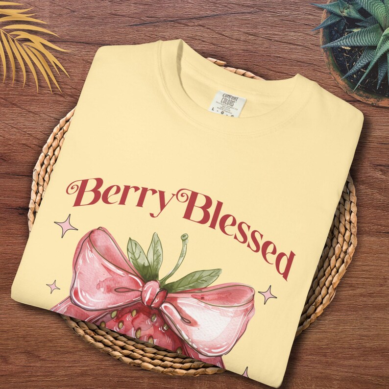 Op de afbeelding: Lichtgeel T-shirt met de tekst "Berry Blessed" in bordeauxrood schrift, vergezeld van een aquarel aardbei illustratie. De aardbei heeft een roze strik en groene bladeren. Het shirt is opgevouwen op een gevlochten mand.