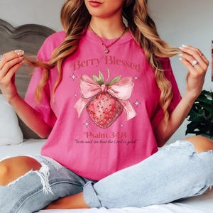 Op de afbeelding: Een roze T-shirt met de tekst "Berry Blessed" boven een aardbei-afbeelding met een roze strik. Daaronder staat de tekst "Psalm 34:8 Proef en zie dat de Heer goed is".