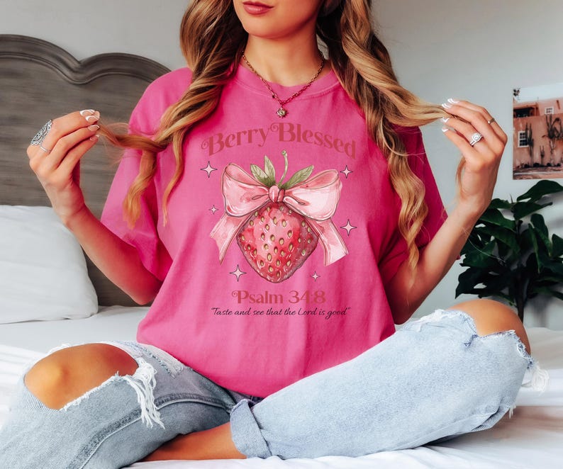 Op de afbeelding: Een roze T-shirt met de tekst "Berry Blessed" boven een aardbei-afbeelding met een roze strik. Daaronder staat de tekst "Psalm 34:8 Proef en zie dat de Heer goed is".