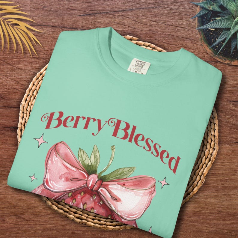 Op de afbeelding: Een lichtgroen T-shirt met de tekst "Berry Blessed" in rode letters. Het shirt heeft een aquarel illustratie van een aardbei met een roze strik en groene bladeren. Het shirt is op een geweven oppervlak gevouwen.