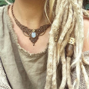 May include: A woman with long blonde dreadlocks wearing a brown crocheted top and a brown macrame necklace with a light blue gemstone pendant.