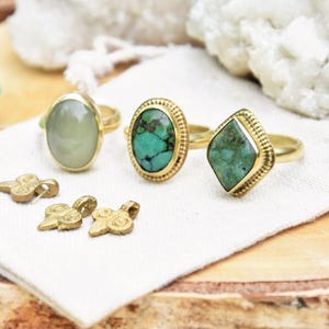 May include: Gold rings with various gemstone settings, including oval and diamond shapes, are displayed on a white cloth. Small gold pendants are also present. The image includes text in Dutch advertising a silversmith workshop.