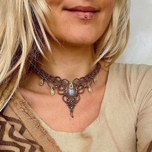 May include: A brown macrame choker necklace with a light blue gemstone pendant and gold accents. The necklace is worn by a person with blonde hair.