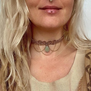 May include: A brown leather choker necklace with a green stone pendant and a chain detail. The necklace is worn by a person with blonde hair.
