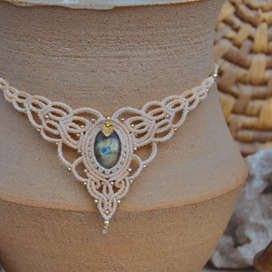 May include: A cream-colored macrame necklace with a central oval stone and gold-colored beads. The necklace is displayed on a beige ceramic vase. The background includes decorative elements like crystals and a woven basket.