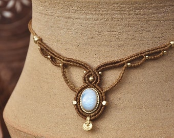 Puede incluir: Un collar gargantilla de macramé marrón con un acento dorado y un colgante de piedra lunar blanca.