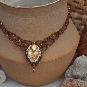 May include: A handcrafted macrame necklace with a central oval-shaped labradorite stone. The necklace features a brown cord with intricate knotting and small gold-colored beads. The labradorite stone has iridescent colors of blue, green, and gold.