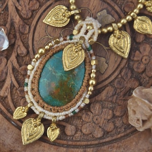 May include: A handcrafted necklace featuring a large, oval turquoise stone pendant. The pendant is framed with macrame cord and accented with gold-colored leaf charms and beads. The necklace rests on a carved wooden surface.
