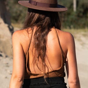 Peut inclure: Une femme portant un haut marron à bretelles et un chapeau fedora marron. Elle se tient dans une zone sablonneuse avec des arbres en arrière-plan.