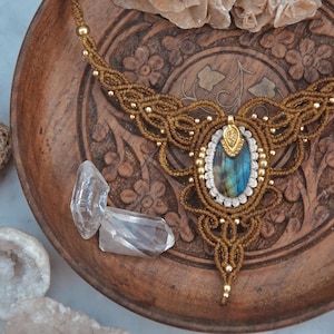 May include: A handcrafted macrame necklace with a central oval-shaped labradorite stone. The necklace features intricate knotting in a warm brown color, accented with gold beads. The necklace is displayed on a carved wooden plate with clear quartz crystals.