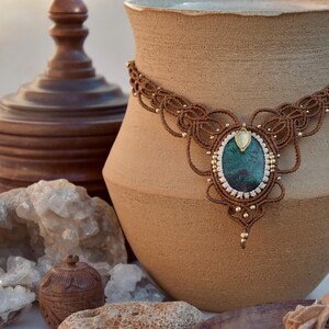 May include: A handcrafted brown macrame necklace with a large oval pendant. The pendant features a teal-colored stone surrounded by white and gold accents. The necklace is displayed on a beige ceramic vase, showcasing bohemian jewelry.