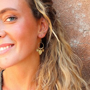 Puede incluir: Una mujer con cabello rubio y ojos azules sonr&iacute;e, usando aretes dorados en forma de hoja. Los aretes tienen un dise&ntilde;o circular con peque&ntilde;as cuentas. El fondo es una pared texturizada de color marr&oacute;n claro.