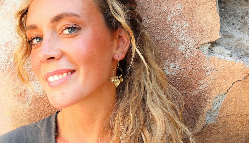 Puede incluir: Una mujer con cabello rubio y ojos azules sonr&iacute;e, usando aretes dorados en forma de hoja. Los aretes tienen un dise&ntilde;o circular con peque&ntilde;as cuentas. El fondo es una pared texturizada de color marr&oacute;n claro.