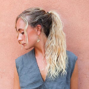 Puede incluir: Una persona con cabello rubio recogido en una cola de caballo lleva pendientes dorados y una blusa sin mangas gris. Los pendientes tienen un dise&ntilde;o &uacute;nico. El fondo es de color melocot&oacute;n s&oacute;lido.