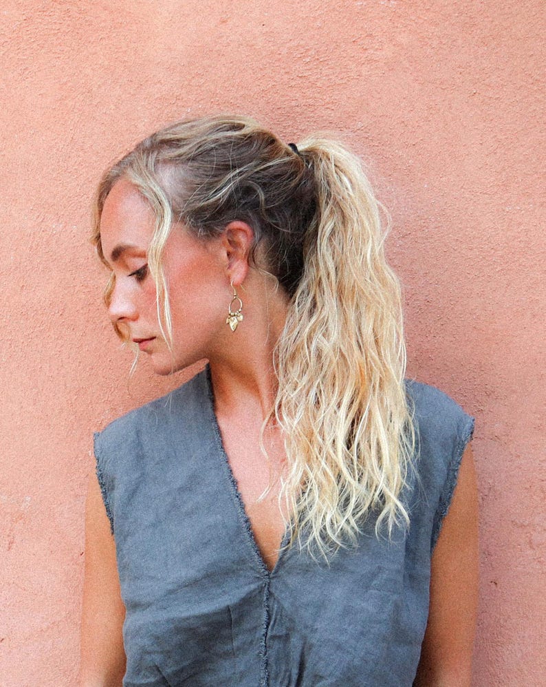 Puede incluir: Una persona con cabello rubio recogido en una cola de caballo lleva pendientes dorados y una blusa sin mangas gris. Los pendientes tienen un dise&ntilde;o &uacute;nico. El fondo es de color melocot&oacute;n s&oacute;lido.