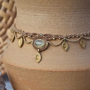 May include: A handcrafted macrame choker necklace with a central oval-shaped stone and dangling gold-colored leaf charms. The necklace features a brown cord and gold-colored beads, creating a bohemian aesthetic. The necklace is displayed against a neutral background.