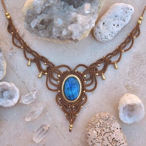 May include: A handcrafted macrame necklace featuring a large, oval blue gemstone set in a gold-colored bezel. The necklace is made with brown cord and accented with small gold beads and teardrop-shaped charms. The design is intricate and bohemian.