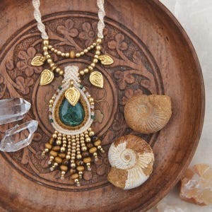 Op de afbeelding: Een handgemaakte macramé ketting met een druppelvormige groene stenen hanger, goudkleurige bladeren en houten kralen. De ketting ligt op een gesneden houten dienblad met twee gefossiliseerde ammonieten.