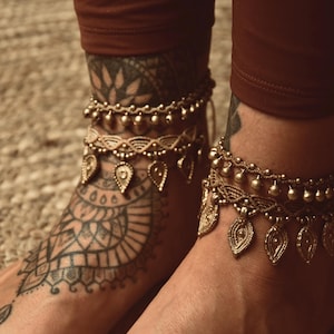 Può includere: Due cavigliere dorate con disegni intricati e ciondoli pendenti. Le cavigliere sono indossate sulle caviglie di una persona, una su ogni caviglia. Le cavigliere sono in metallo e hanno un design a perline.