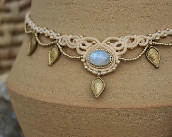 Puede incluir: Un delicado collar de macramé blanco con un gran colgante ovalado de piedra lunar y detalles de hojas doradas.