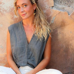 Puede incluir: Una mujer con una blusa gris sin mangas con cuello en V y pantalones color crema. Est&aacute; sentada frente a una pared texturizada de color marr&oacute;n rojizo. Tiene el pelo rubio y lleva pendientes dorados.