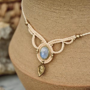 May include: A delicate white macrame necklace with a light blue moonstone pendant and a gold leaf charm. The necklace is made with small gold beads and has a delicate, bohemian style.