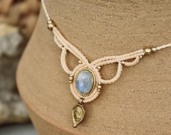 Puede incluir: Un delicado collar de macramé blanco con un colgante de piedra de luna azul claro y un dije de hoja de oro. El collar está hecho con pequeñas cuentas doradas y tiene un estilo delicado y bohemio.