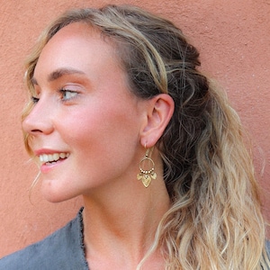 Puede incluir: Pendientes colgantes dorados con una parte superior circular y una parte inferior decorativa en forma de hoja. Los pendientes los lleva una persona con cabello rubio y una parte superior gris, sobre un fondo color melocot&oacute;n.