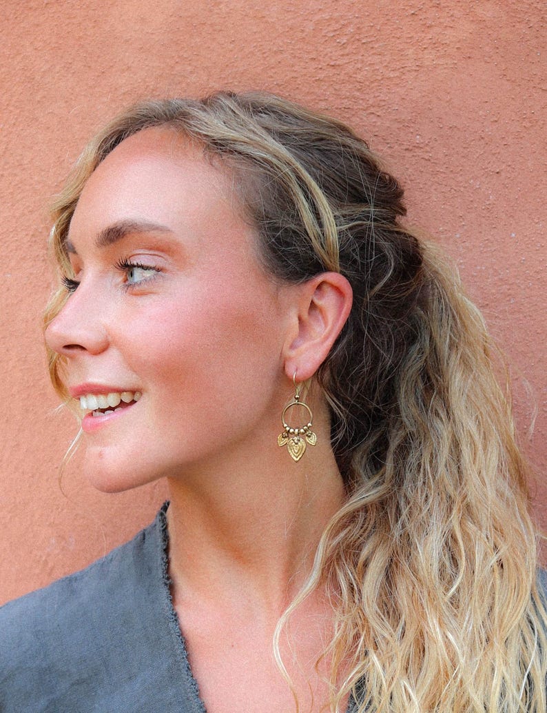 Puede incluir: Pendientes colgantes dorados con una parte superior circular y una parte inferior decorativa en forma de hoja. Los pendientes los lleva una persona con cabello rubio y una parte superior gris, sobre un fondo color melocot&oacute;n.