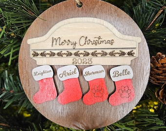 May include: A round wooden Christmas ornament with a light brown border and a darker brown center. It features the words "Merry Christmas 2023" and four red stockings with names and paw prints. A jute string is attached for hanging.
