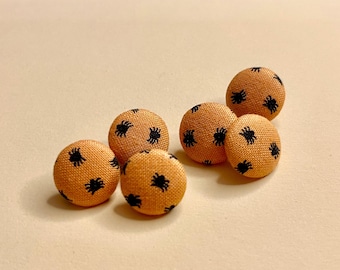 May include: A set of five round, fabric-covered buttons featuring a textured orange surface with a repeating pattern of small, black spider icons printed across each button. The buttons have a domed shape and a consistent woven textile appearance.