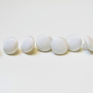 May include: A selection of white, fabric-covered buttons with a textured weave. The buttons are round and have a small, clear plastic shank on the back. The buttons are arranged in a row against a white background.
