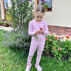 May include: A child in a light pink long-sleeved top and trousers, holding a red flower. The child is wearing light pink clogs. The background features green grass, colourful flowers, and a building with a window.