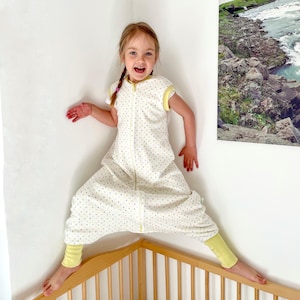 May include: A cream-colored sleep sack with a polka dot pattern, yellow trim, and a full-length zipper. The sleep sack is worn by a young child. The child is wearing yellow leggings and has their arms outstretched.