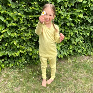 May include: A young person wearing a light yellow long-sleeved shirt and matching pants, holding a daisy. The outfit is a solid color, and the person is standing on grass in front of a green hedge.