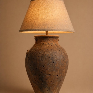May include: A rustic table lamp with a textured, brown ceramic base featuring numerous small holes. The lamp has a beige linen lampshade. The lamp is designed to provide warm, ambient lighting.