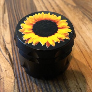 Pu&ograve; includere: Macinino in metallo nero con un design a girasole sulla parte superiore. Il girasole ha petali gialli e un centro marrone scuro.