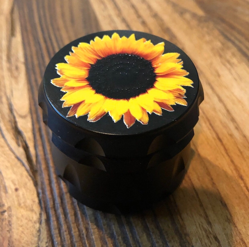 Pu&ograve; includere: Macinino in metallo nero con un design a girasole sulla parte superiore. Il girasole ha petali gialli e un centro marrone scuro.