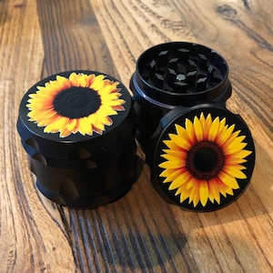 Pu&ograve; includere: Macinino in metallo nero con tre sezioni. Ogni sezione presenta un design a girasole con petali gialli e un centro marrone.